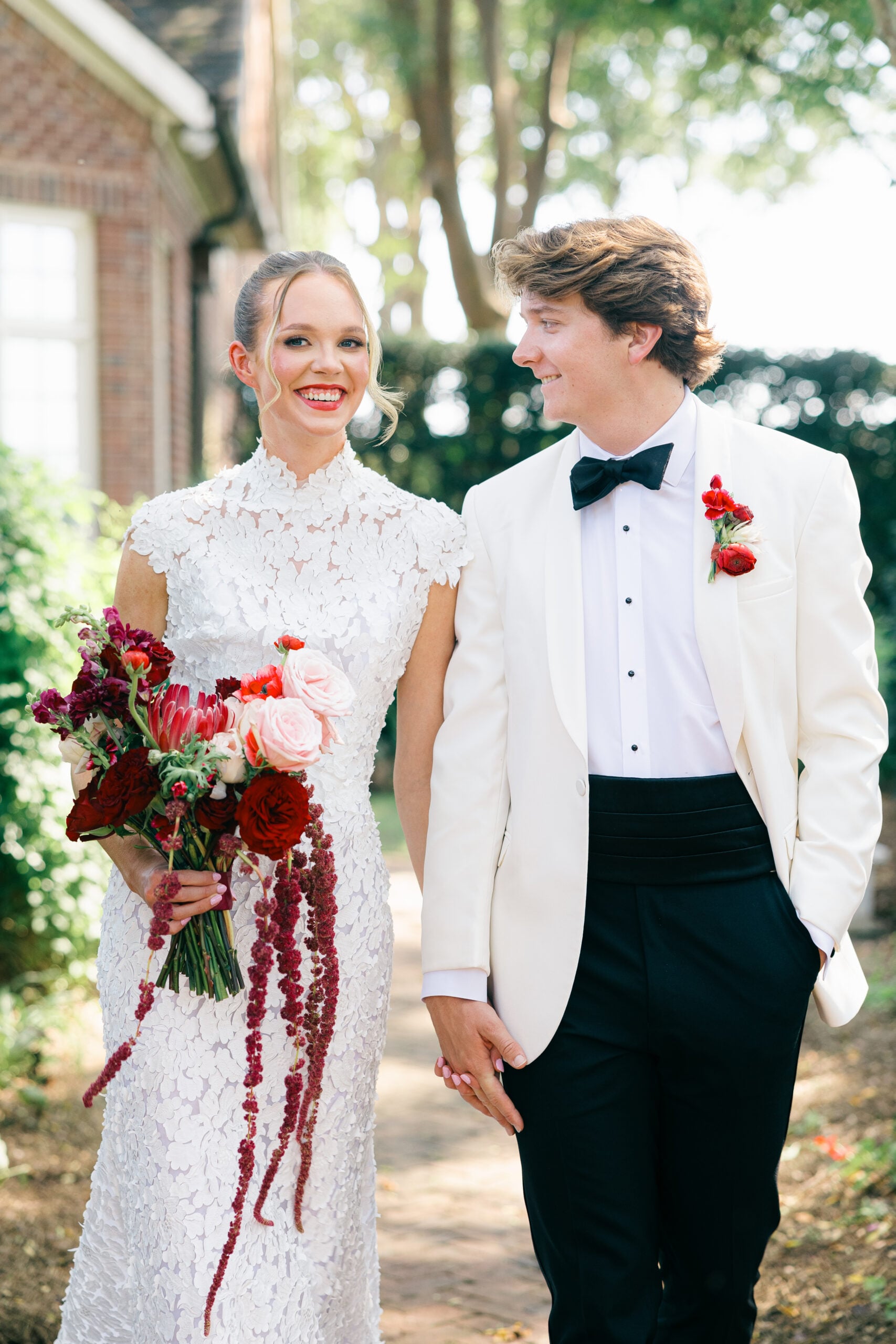 A bride in a white lace gown and a groom in a white dinner jacket walk hand in hand, holding a bouquet of red and pink flowers.